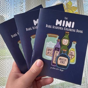 Op de afbeelding: Drie donkerblauwe kleurboeken getiteld The Mini Dark Academia Coloring Book. De kaft bevat illustraties van glazen potten met diverse inhoud, gelabeld met Arrowroot, Belladonna, Beetle Eyes en Calendula, en de tekst serafine art & design.