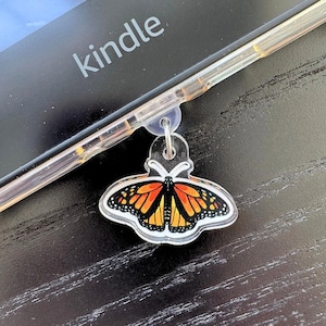 May include: A clear acrylic charm featuring a monarch butterfly design with orange, black, and yellow wings. The charm is attached to a clear phone case. The word "kindle" is visible on the background.