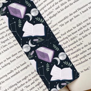 May include: A bookmark with a dark blue background featuring illustrations of open and closed books, moons, constellations, and sprigs of lavender. The bookmark has a whimsical, celestial theme.