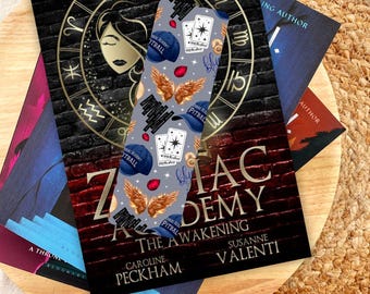 May include: A gray and blue bookmark with a repeating pattern of playing cards, wings, and the words "Zodiac Academy" and "Pitball".