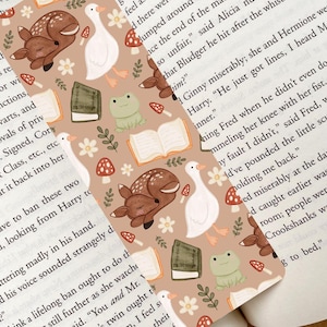 May include: A whimsical bookmark featuring a repeating pattern of a sleeping deer, white goose, frog, mushrooms, books, and flowers on a tan background. The bookmark is rectangular and made of paper.