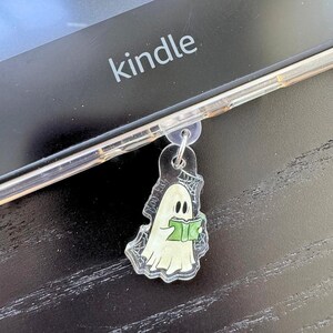May include: A clear acrylic charm featuring a cartoon ghost reading a green book, with a spiderweb design. The charm is attached to a clear phone case, resting on a black surface with the word "kindle" visible.