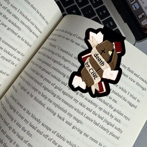 May include: A sticker featuring a brown coffin with a cross, a banner reading "death by CBR", and a black outline. The sticker is placed on an open book with printed text.