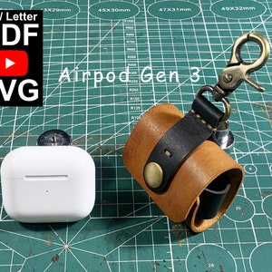 May include: A brown leather case with a black leather strap and a brass hook. The case is designed to hold a white Airpod Gen 3 case. The case is sitting on a green cutting mat with white markings.