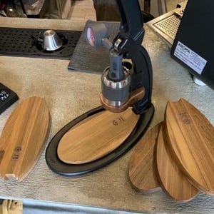 Puede incluir: Una cafetera espresso manual negra y plateada con base y mango de madera. Varias tablas de servir de madera están dispuestas alrededor de la cafetera. Las tablas tienen forma ovalada y un acabado marrón claro.