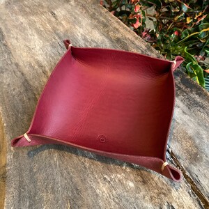May include: A burgundy leather catchall tray with a square shape and curved sides. The corners are secured with light-colored stitching. The tray has a small circular logo on the bottom.