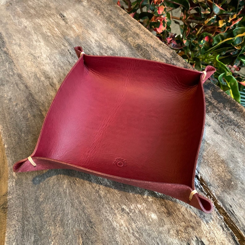 May include: A burgundy leather catchall tray with a square shape and curved sides. The corners are secured with light-colored stitching. The tray has a small circular logo on the bottom.