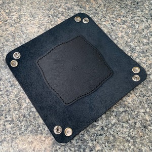 May include: A black leather valet tray with a square shape and a smaller, textured square in the center. The tray has metal snap closures at each corner. The tray is designed to hold small items like keys or wallets.