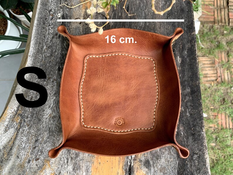 May include: A small, square, brown leather tray with a stitched border and a button detail. The tray measures 6.3 inches across. The letter "S" is visible in the image. The tray is designed to hold small items.