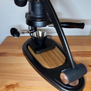 May include: A black Flair espresso maker with a wooden base and a stainless steel portafilter. The machine has a pressure gauge and a lever for brewing espresso.