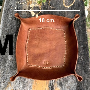 May include: A brown leather tray, approximately 7 inches wide, with a stitched border and a central, lighter-colored square. The tray has curved edges and a button detail. The letter "M" is visible in the image.
