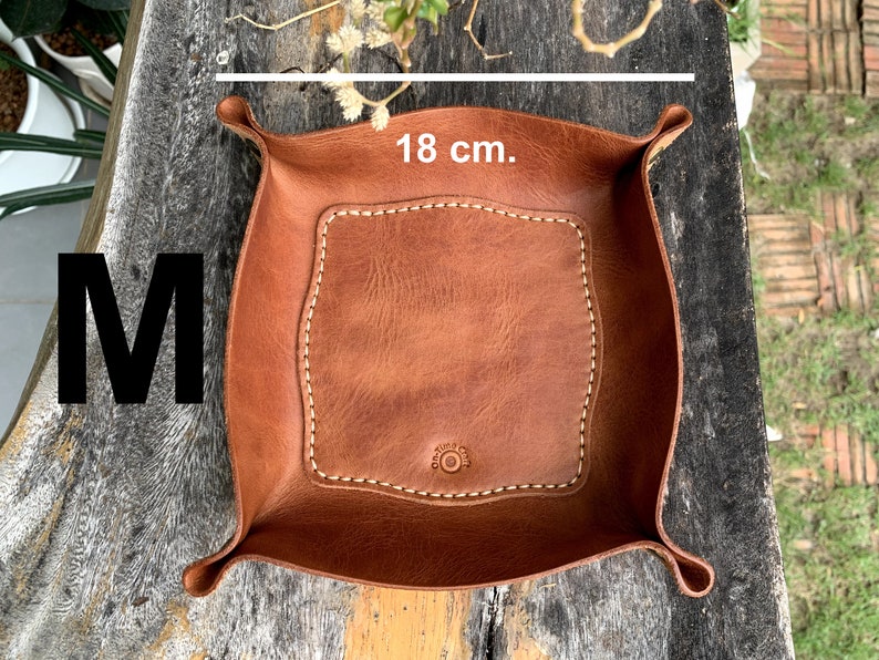 May include: A brown leather tray, approximately 7 inches wide, with a stitched border and a central, lighter-colored square. The tray has curved edges and a button detail. The letter "M" is visible in the image.