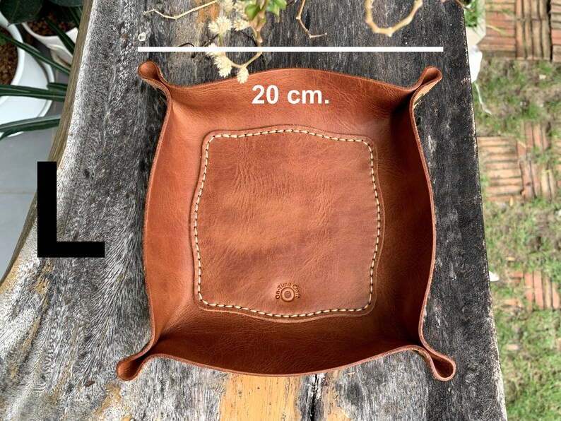 May include: A brown leather tray with a stitched border and a central, lighter-colored square. The tray is approximately 7.9 inches wide and has curved edges with raised corners. The letter "L" is in the image.