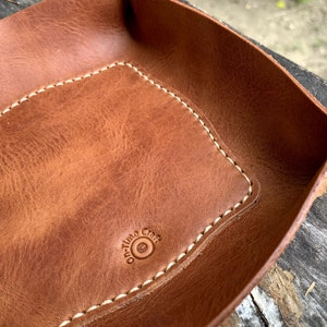 May include: A brown leather catchall tray with a rectangular interior and rounded corners. The tray features white stitching around the edges and a stamped logo that reads "On-time Craft". The tray has a snap closure.