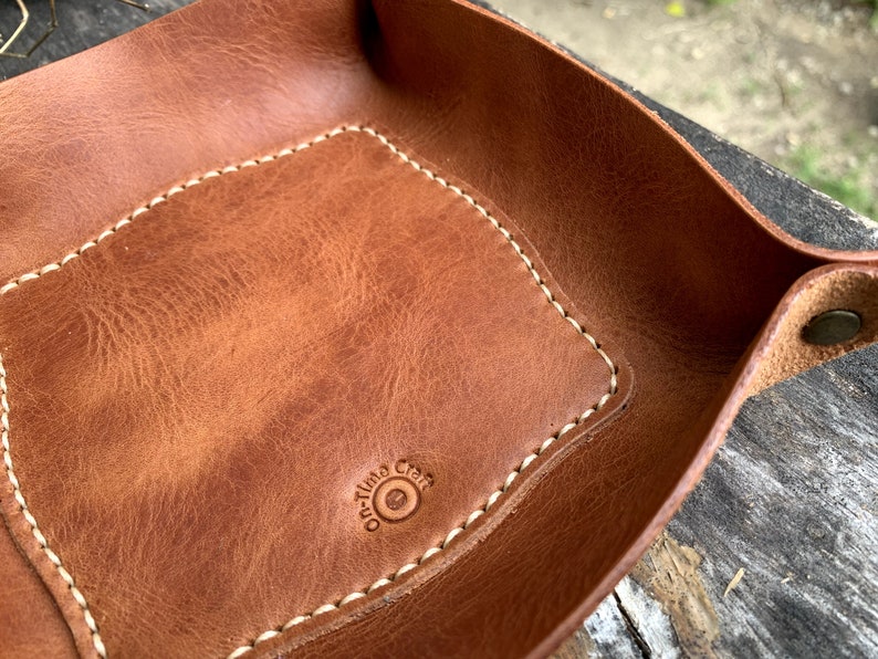May include: A brown leather catchall tray with a rectangular interior and rounded corners. The tray features white stitching around the edges and a stamped logo that reads "On-time Craft". The tray has a snap closure.