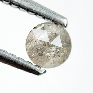 Op de afbeelding: Een ronde, lichtgrijze diamant, vastgehouden door een zilveren pincet. De diamant heeft een gefacetteerd oppervlak met zichtbare interne insluitsels. De pincet is schuin geplaatst, waardoor de edelsteen tegen een witte achtergrond wordt getoond.