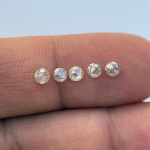 Puede incluir: Cinco pequeños diamantes redondos y claros están dispuestos en fila. Los diamantes tienen una superficie facetada y están colocados sobre un fondo borroso. Los diamantes se sostienen en el dedo de una persona.