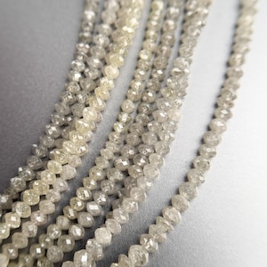 Peut inclure: Gros plan sur plusieurs brins de perles de diamants bruts. Les perles sont un mélange de clair et de gris, avec une forme facettée et irrégulière. Les brins sont disposés en cascade sur un fond neutre.