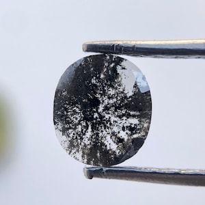 Op de afbeelding: Een ronde, heldere edelsteen met zwarte insluitsels, vastgehouden door een pincet. De insluitsels vormen een uniek, boomachtig patroon in de steen. De edelsteen is gepolijst en heeft een reflecterend oppervlak.