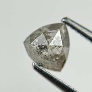 Puede incluir: Un diamante gris en forma de triángulo, sujeto con unas pinzas. El diamante tiene una superficie facetada con inclusiones internas y un aspecto ligeramente translúcido. Las pinzas son de color gris oscuro.