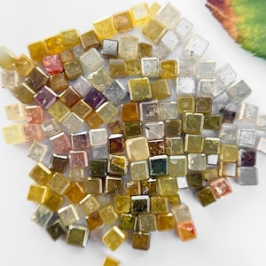Pode incluir: Uma coleção de pequenos diamantes quadrados facetados em vários tons de amarelo, marrom e verde. Os diamantes estão soltos e espalhados sobre uma superfície branca.