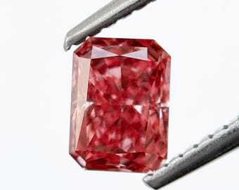 Op de afbeelding: Een stralende, rechthoekig geslepen, fancy rode diamant, vastgehouden door een zilveren pincet. De edelsteen vertoont een complexe interne structuur, die licht reflecteert en een sprankelend effect creëert. De kleur en slijpvorm van de diamant worden benadrukt tegen een witte achtergrond.