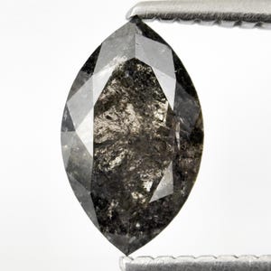 May include: A marquise-cut, black diamond gemstone held by a metal tool. The diamond has a faceted surface with internal inclusions, displaying a mix of dark gray and black tones. The gemstone is set against a white background.
