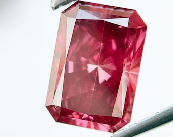 Op de afbeelding: Een rechthoekig geslepen, roze edelsteen, vastgehouden door een pincet. De edelsteen heeft een complexe interne structuur met meerdere facetten die het licht reflecteren en een sterpatroon creëren. De kleur van de steen is diep, verzadigd roze.