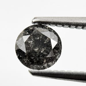 Op de afbeelding: Een ronde, gefacetteerde edelsteen in donkergrijs tot zwart met een gespikkelde, ondoorzichtige uitstraling. De steen wordt vastgehouden door een zilverkleurige metalen pincet, wat de ronde slijpvorm en textuur met zichtbare insluitingen benadrukt.