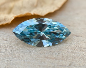 May include: A marquise-cut blue gemstone, with a faceted design. The gemstone is displayed against a light brown background.