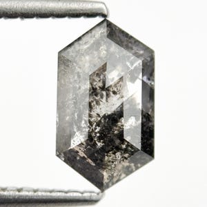 May include: A hexagonal-shaped, salt and pepper diamond with a clear, gray body and dark inclusions. The diamond is held by a metal tool against a white background. The facets of the diamond are visible.
