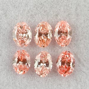 Puede incluir: Seis piedras preciosas de corte ovalado con un tono rosa melocotón claro y superficies facetadas, dispuestas en dos filas de tres. Cada piedra muestra una calidad brillante y reflectante con cortes simétricos que crean destellos.