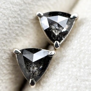 Könnte beinhalten: Ein Paar dreieckig geschliffene, schwarze Diamant-Ohrstecker. Jeder Ohrring hat einen dunklen, facettierten Diamanten, der von drei silbernen Zinken gehalten wird. Die Diamanten haben ein gesprenkeltes Aussehen und reflektieren das Licht.