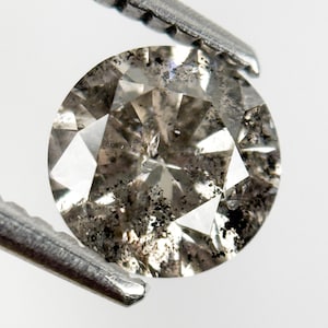 May include: A round, salt and pepper diamond held by a metal tool. The diamond has a faceted cut with a mix of clear and dark inclusions. The stone is a neutral color, with a speckled appearance.
