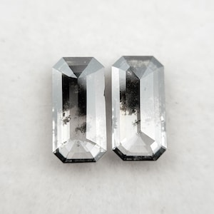 Può includere: Due diamanti sale e pepe a taglio smeraldo rettangolare con superficie sfaccettata, inclusioni scure visibili e toni grigio-bianchi. Queste pietre preziose presentano una finitura lucida e chiara con bordi geometrici ben definiti.