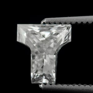 Può includere: Una pietra preziosa trasparente e incolore tagliata a forma di T con molteplici sfaccettature che riflettono la luce. La pietra è tenuta da una pinzetta metallica, che mette in mostra il suo design geometrico e la struttura cristallina.