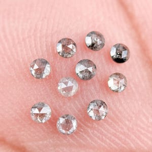 May include: Nine round, rose-cut diamonds in varying shades of grey and clear. The gems, with visible inclusions, are displayed against a pink background. Each diamond's facets are visible, highlighting their sparkle.