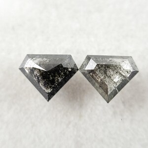 May include: Two shield-cut loose diamonds featuring a salt and pepper appearance with dark grey and black inclusions. The stones have a faceted surface with a geometric shape, displaying a mix of opaque dark areas and translucent grey sections.