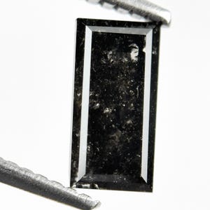 May include: A rectangular, black gemstone with a white border. The gemstone has a speckled appearance and is held by a metal tool. The gem is set against a white background.