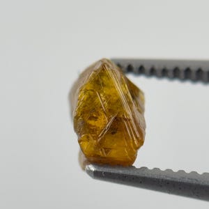 May include: A raw, translucent amber-coloured gemstone with a multifaceted, irregular crystalline structure. The stone exhibits deep golden-brown hues and sharp, geometric edges, held securely by a pair of metallic grey serrated tweezers.
