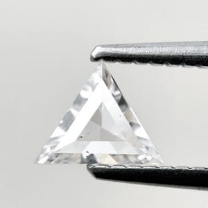 Könnte beinhalten: Ein klarer, dreieckig geschliffener Edelstein, der von einer Pinzette gehalten wird. Der Edelstein hat mehrere Facetten, die das Licht reflektieren. Die Form ist ein gleichseitiges Dreieck. Die Pinzette ist silberfarben.