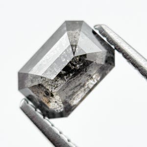 Op de afbeelding: Een rechthoekig geslepen diamant met een zout-en-peper-uiterlijk, vastgehouden door een zilveren juweliersgereedschap. De edelsteen heeft grijze en zwarte insluitsels, tegen een witte achtergrond.