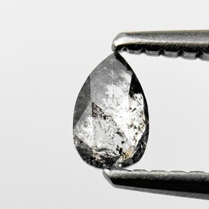 May include: A pear-shaped, salt and pepper diamond held by a pair of tweezers. The diamond has a mix of gray and white inclusions, with a polished surface. The tweezers are silver-colored, and the background is white.