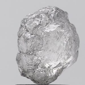 Pode incluir: Um diamante bruto, não lapidado, com uma estrutura cristalina. A gema é de cor cinza claro e tem uma superfície texturizada com facetas visíveis. O diamante é exibido sobre um fundo liso e claro.