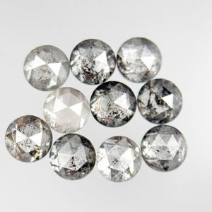 May include: Ten round, faceted diamonds are displayed. The gems are mostly grey and clear, with some having dark inclusions. The facets reflect light, producing a sparkling effect. The diamonds are arranged on a white surface.