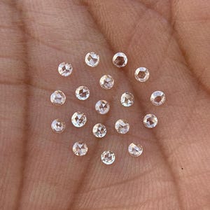 May include: A collection of 20 round, clear, faceted, rose cut diamonds. The diamonds are arranged in a circular pattern on a brown surface.