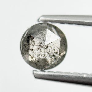May include: A round, gray diamond with a faceted surface is held by silver tweezers. The diamond has a speckled appearance with dark inclusions and a polished, reflective surface. The background is plain white.