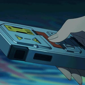 May include: A rectangular, silver-gray device with yellow and red accents. The device has a series of buttons and indicators, and is held in a hand. The background is a blurred blue and teal.