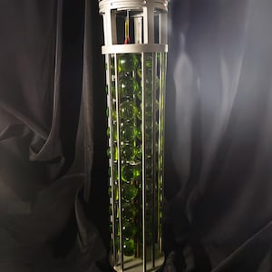 Op de afbeelding: Een hoge, cilindrische kooi van zilverkleurig metaal met een kolom van groene glazen knikkers erin. De kooi heeft een decoratief bovenstuk.