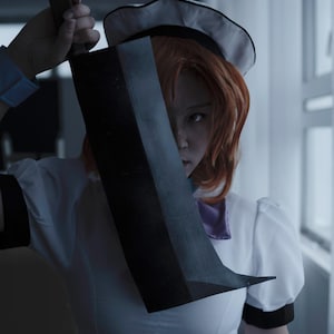 May include: A woman in a white sailor dress holds a large black knife in front of her face. She is looking at the camera with a serious expression.